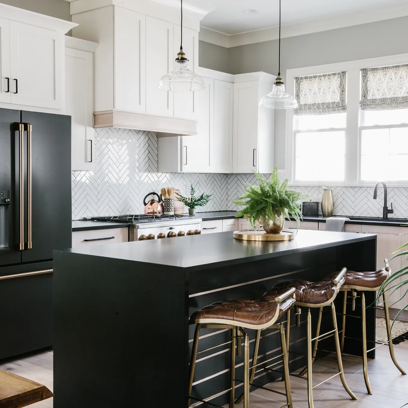 a standout waterfall quartz kitchen island made from Cambria Blackpool Matte is in the center of a white kitchen with plenty of cabinets and natural lighting, a black fridge, and three leather bar stools. 