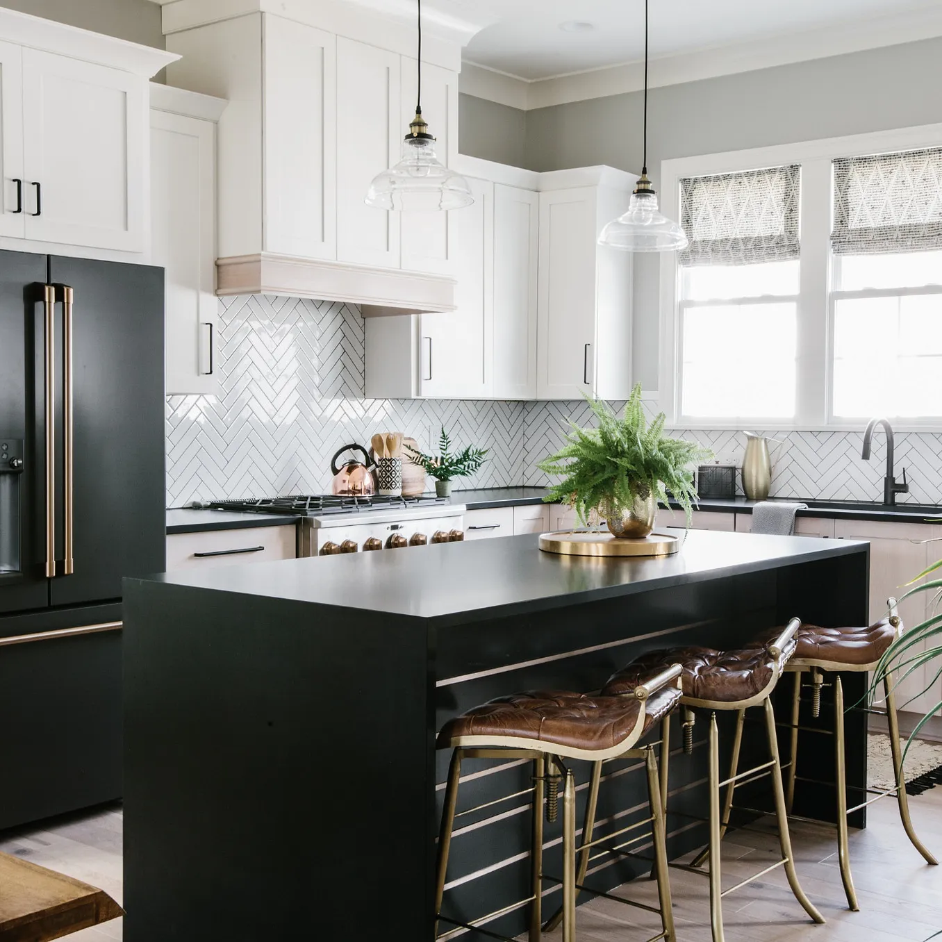 a standout waterfall quartz kitchen island made from Cambria Blackpool Matte is in the center of a white kitchen with plenty of cabinets and natural lighting, a black fridge, and three leather bar stools.