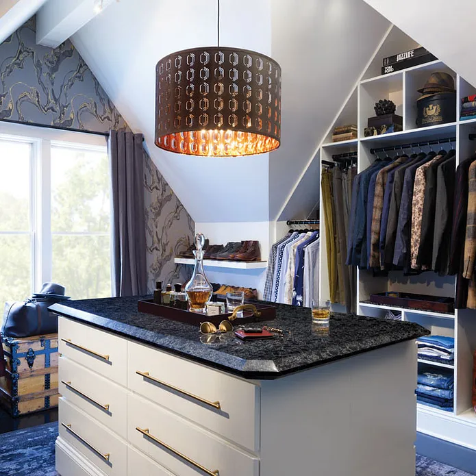 a men's closet with an island featuring Cambria Sharpham quartz countertops, a large chandelier over the island, blue carpet and patterned wallpaper, and open shelving to hang clothes. 