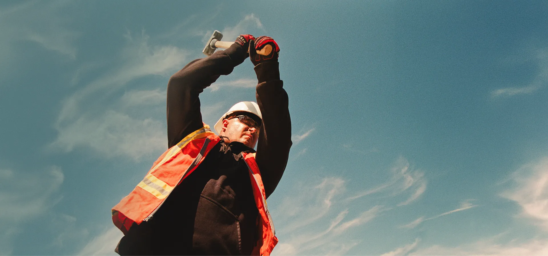 A man in workwear swinging a mallet with a blue sky in the background