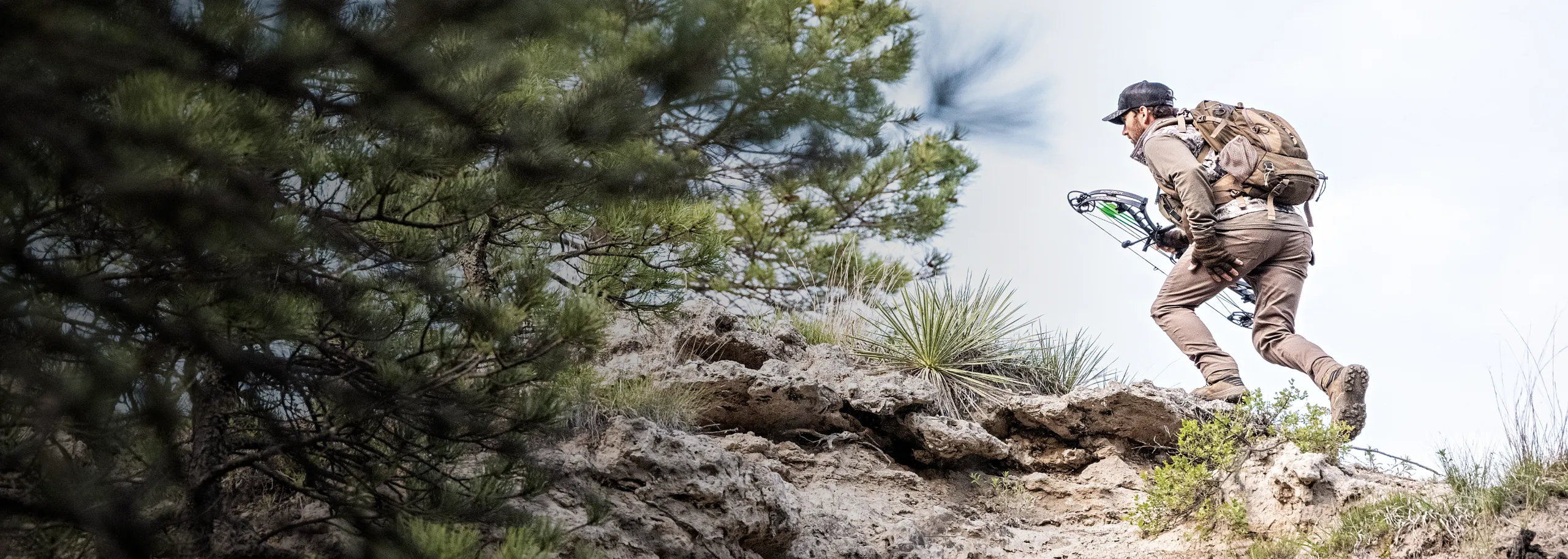 A man in hunting gear with a bow running up a rocky hill