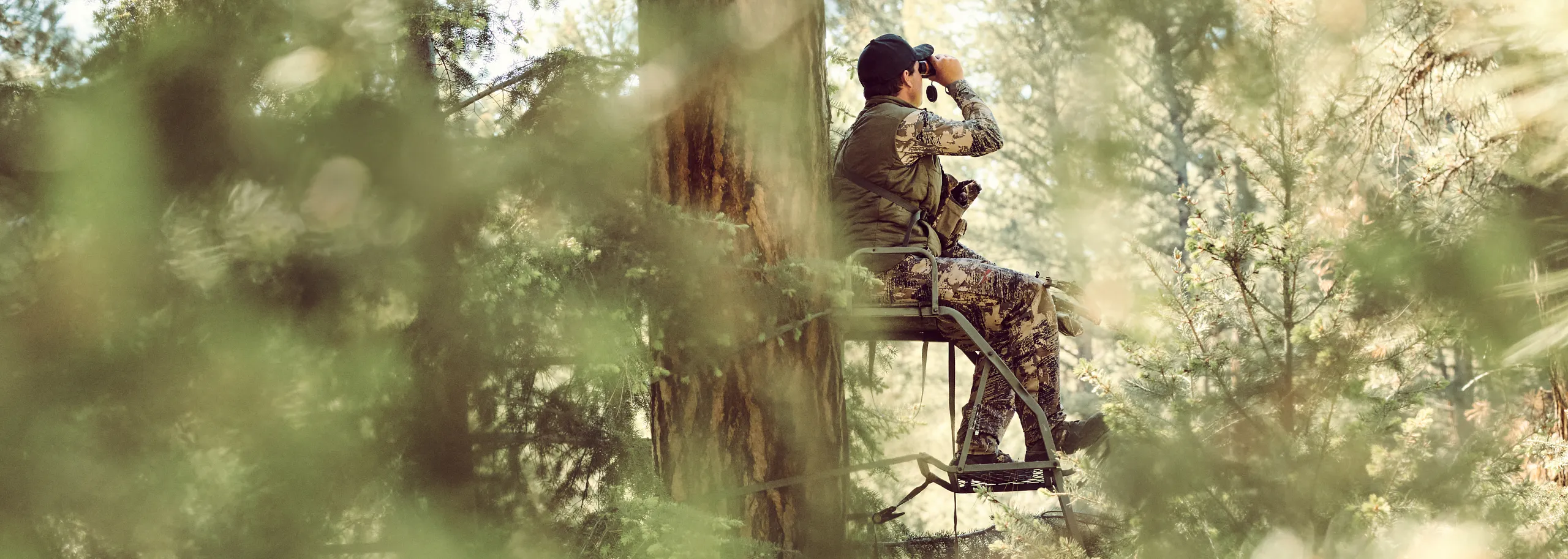 Person sitting in a deer stand in the woods using binoculars wearing Vaprtrek boots