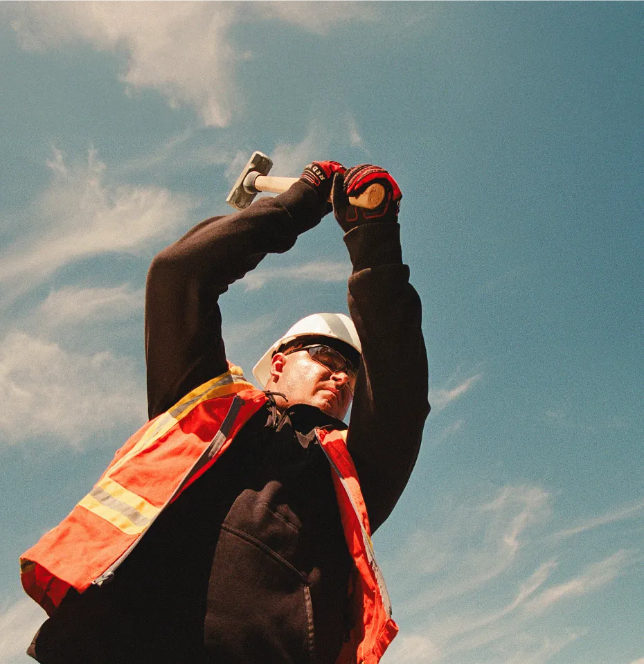 A man in workwear swinging a mallet with a blue sky in the background