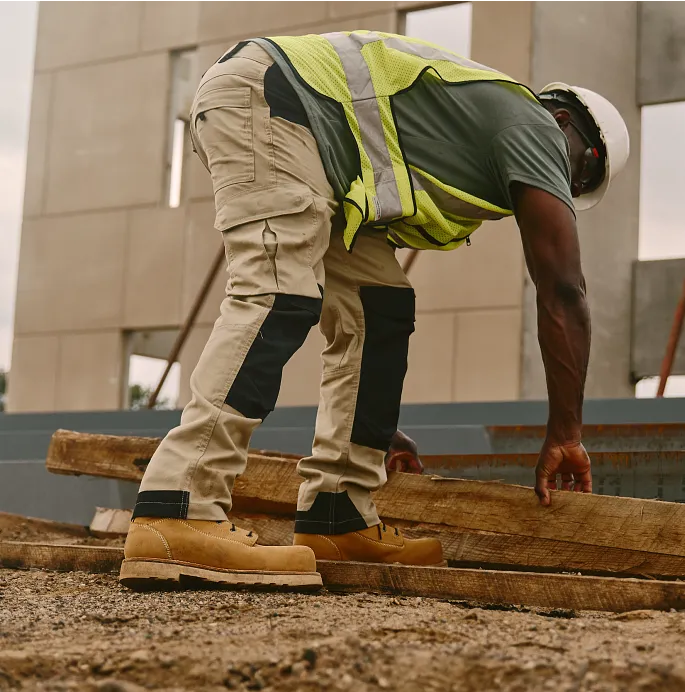 A man in workwear at a worksite bending over to pick up boards