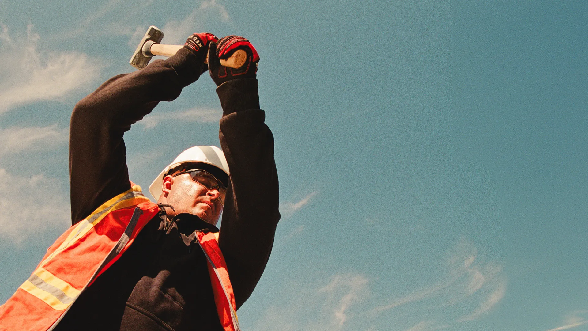 A man in workwear swinging a sledgehammer with a blue sky behind him