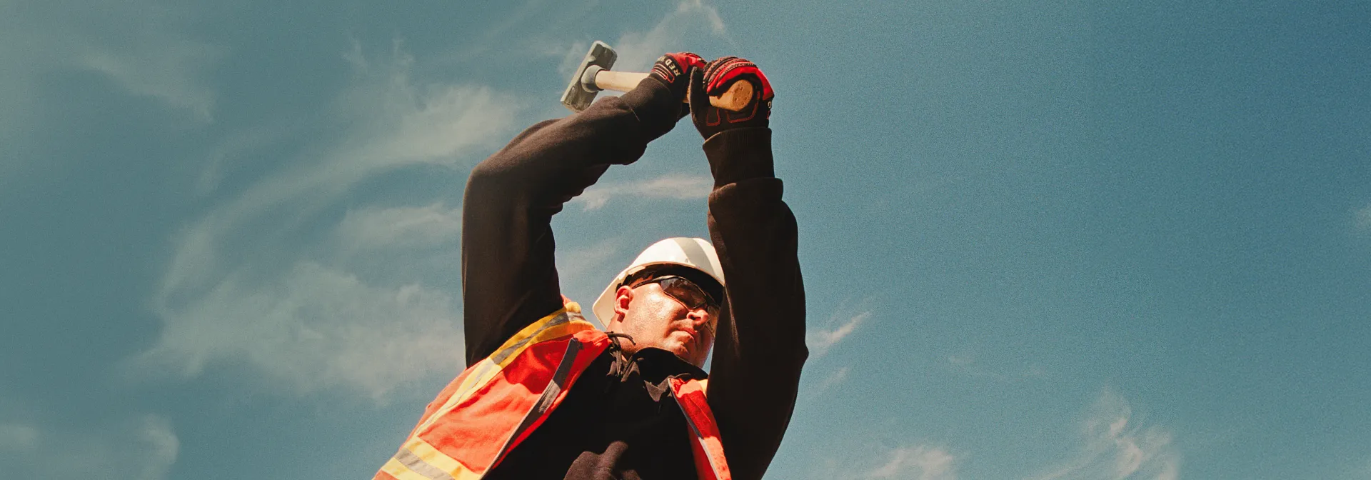 A man in workwear swinging a mallet with a blue sky in the background
