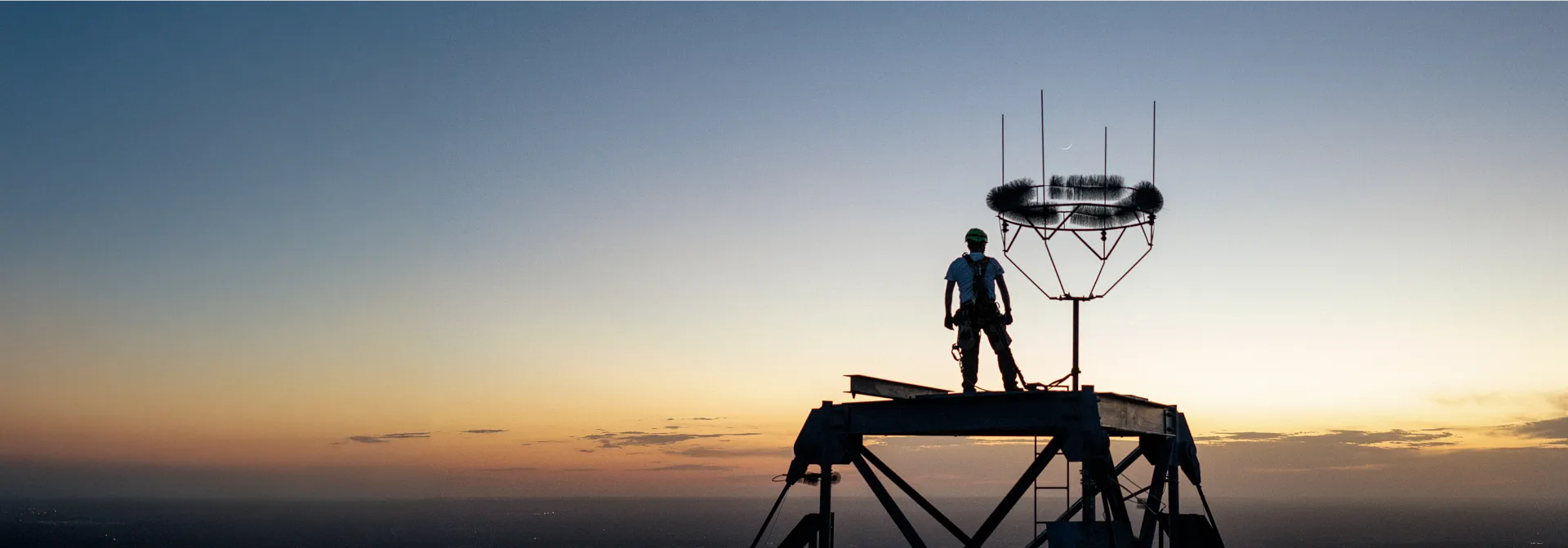 A man standing at the top of a tall structure with a clear sky at dusk in the background