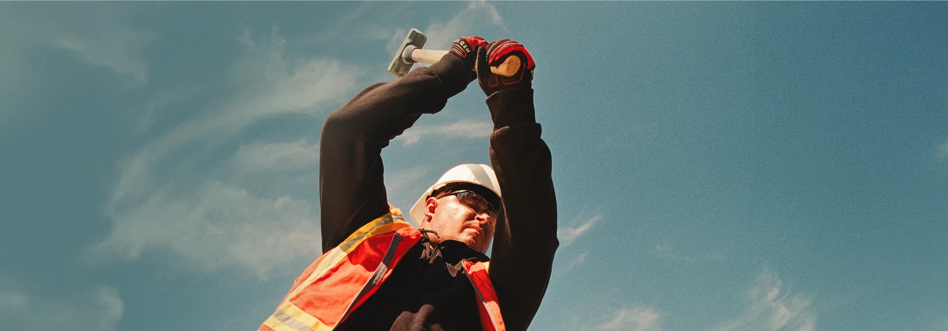 A man in workwear swinging a mallet with a blue sky in the background