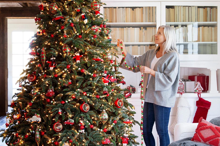 Frau, die farbenfrohe Girlanden an einen geschmückten künstlichen Weihnachtsbaum hängt.