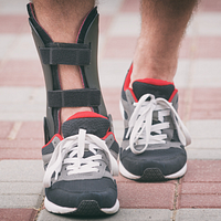 Feet walking in sneakers, one wearing an ankle orthotic brace.