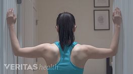 Woman with her back facing the camera and both forearms on opposite walls in preparation to perform the corner stretch