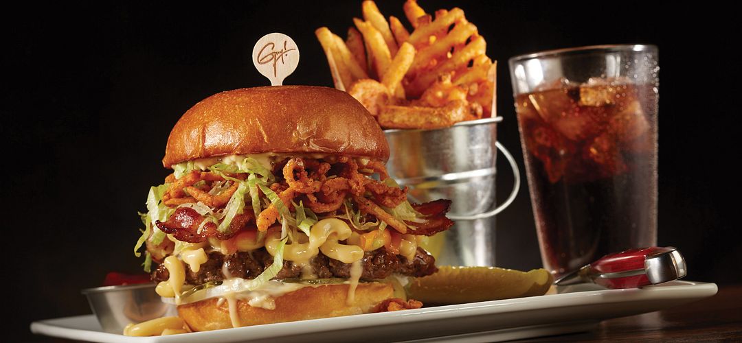 Planet Hollywood Guy Fieri’s Bacon Mac-n-Cheese Big Bite Burger, with fries and a drink in the background, at Planet Hollywood