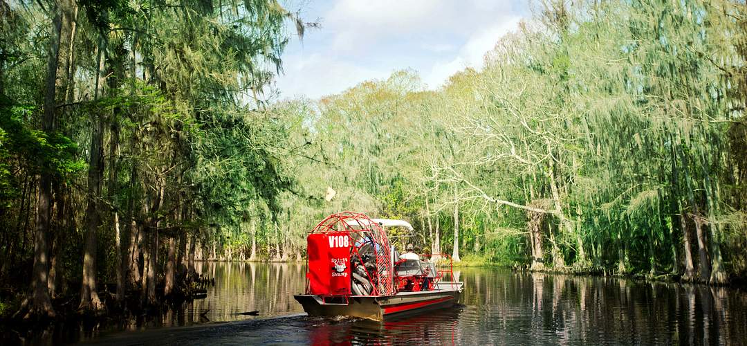 Spirit of the Swamp Airboat Rides Spirit of the Swamp Airboat Rides carries passengers on a red airboat as it glides over calm waters surrounded by cypress trees.