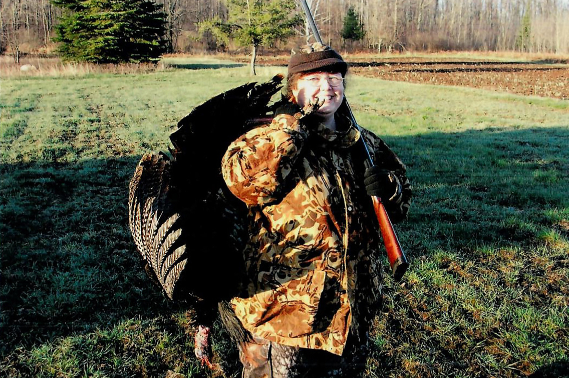 Woman in camouflage clothing in outdoor setting, holding turkey over her shoulder