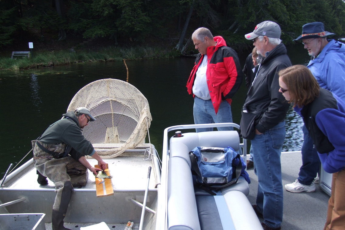 Several people looking on while a man in a boat takes a water sample