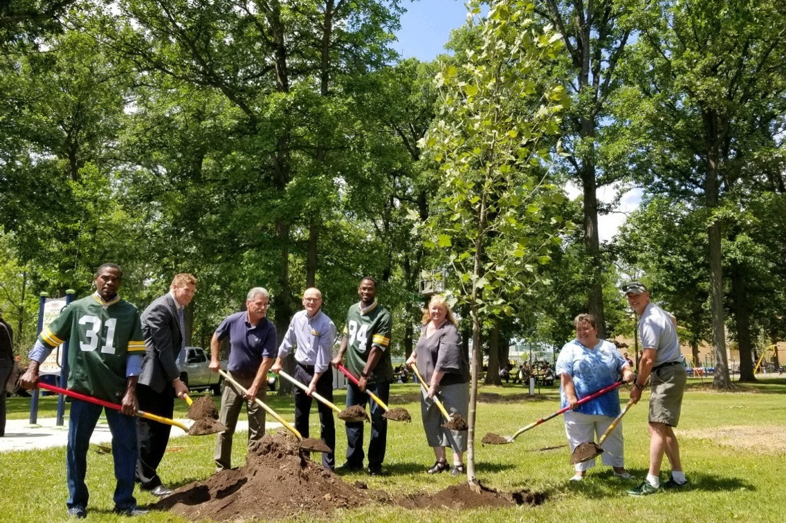 group of people holding shovels and helping plant a tree in a city park