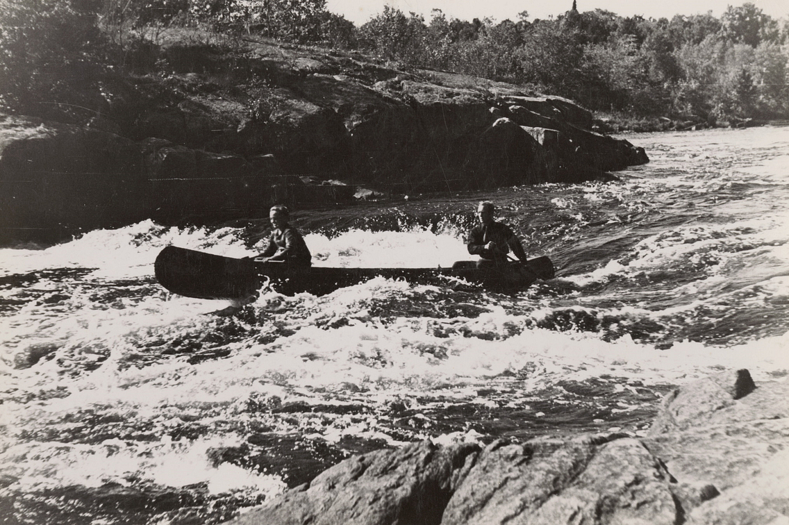 Vintage black and white photo of two men paddling canoe through river rapids