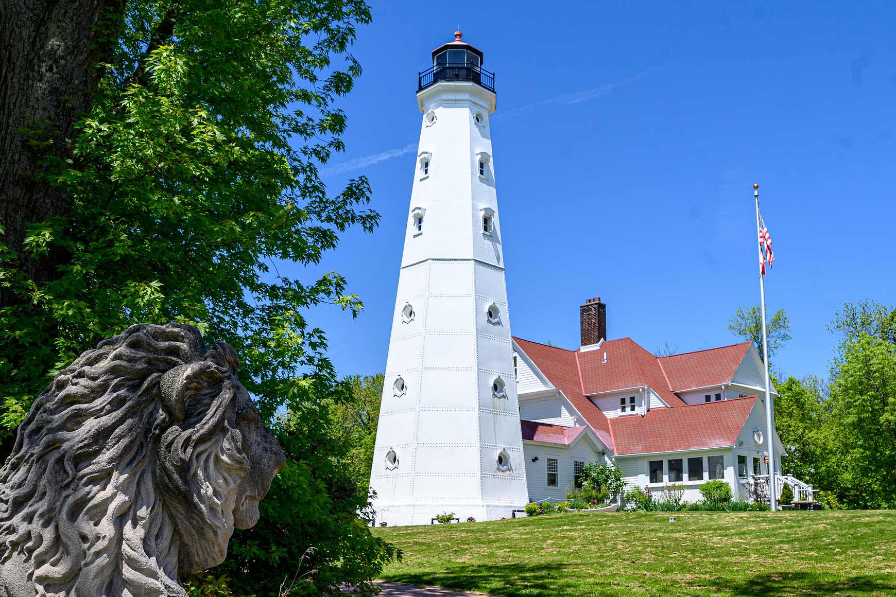 Photo of tall white lighthouse with red-roofed building against clear blue sky and statue of lion in foreground