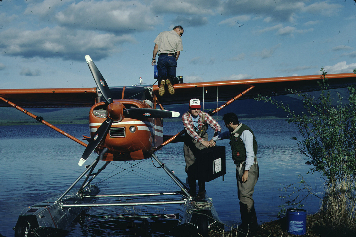 Three men working beside a seaplane in the water, one kneeling on the wings and the other two exchanging a suitcase