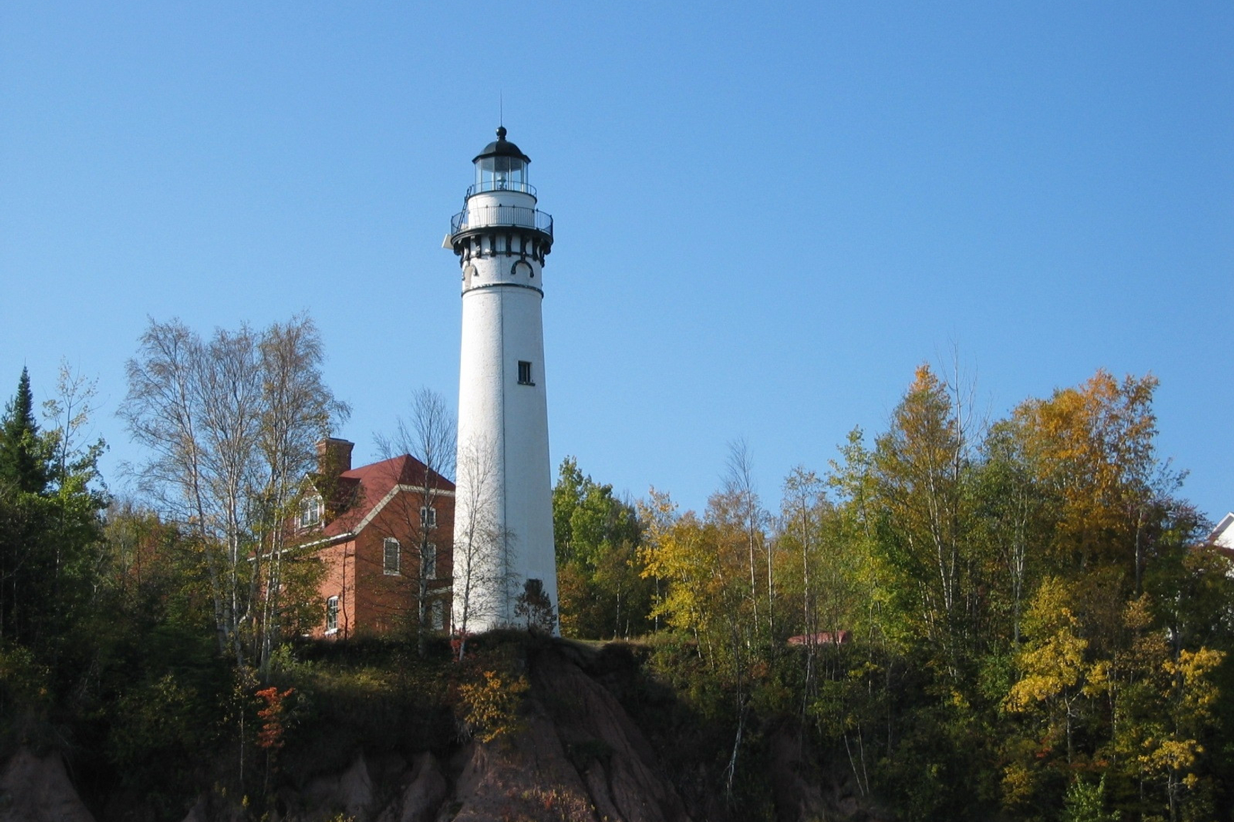Tall white lighthouse stands next to red brick building in forested landscape against clear blue sky