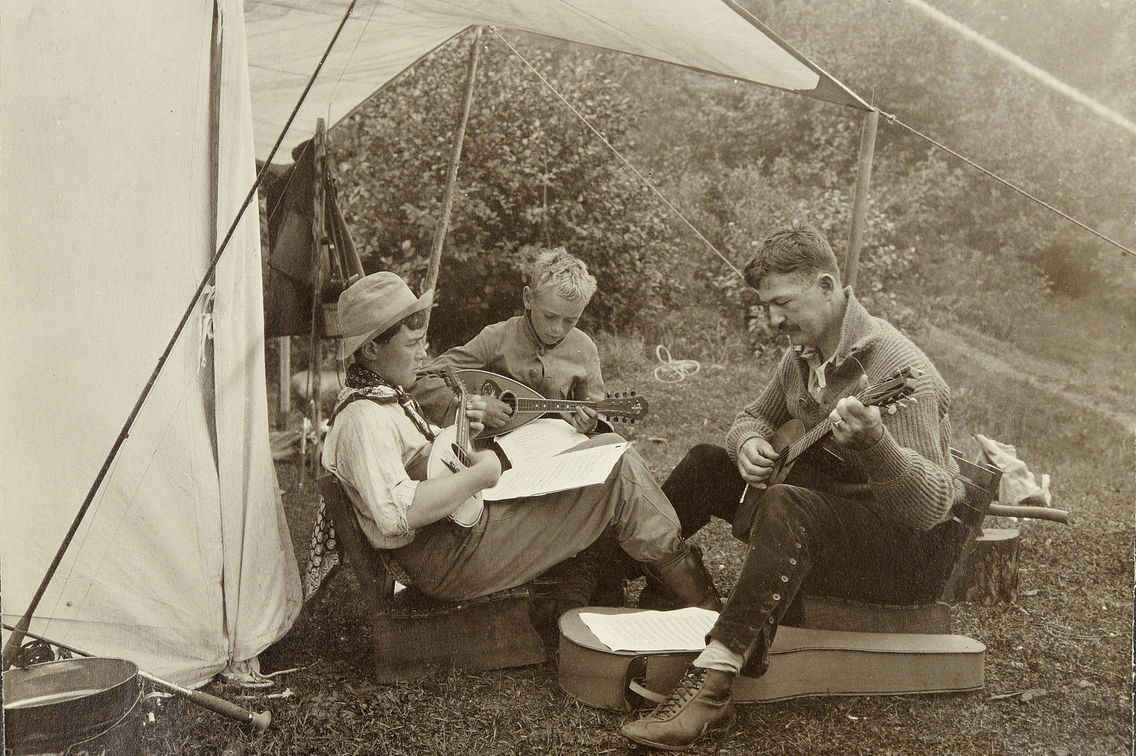 Old photo of three boys sitting under tent flap, one playing a mandolin