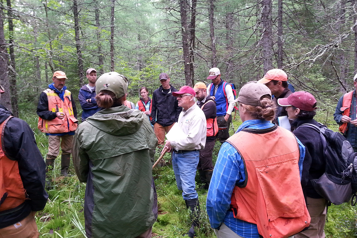 Man in red hat talking among a group of people standing in the forest