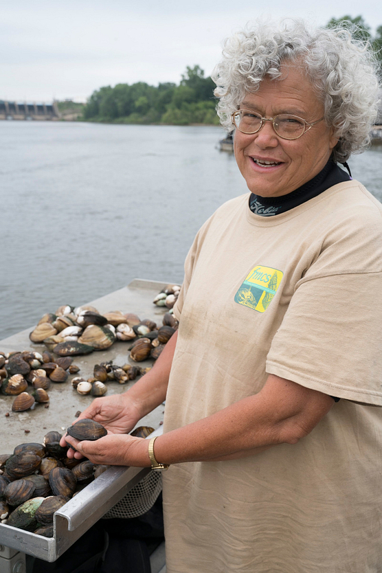 Woman with gray curly hair, wearing a beige T-shirt with logo, holding a mussell in her hands