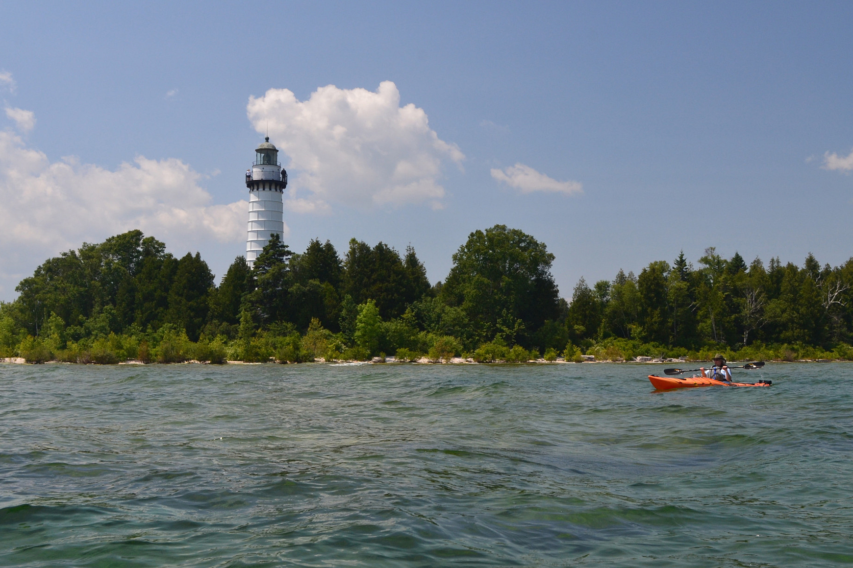 Photo of tall white lighthouse on island in Lake Michigan with kayakers paddling in water nearby