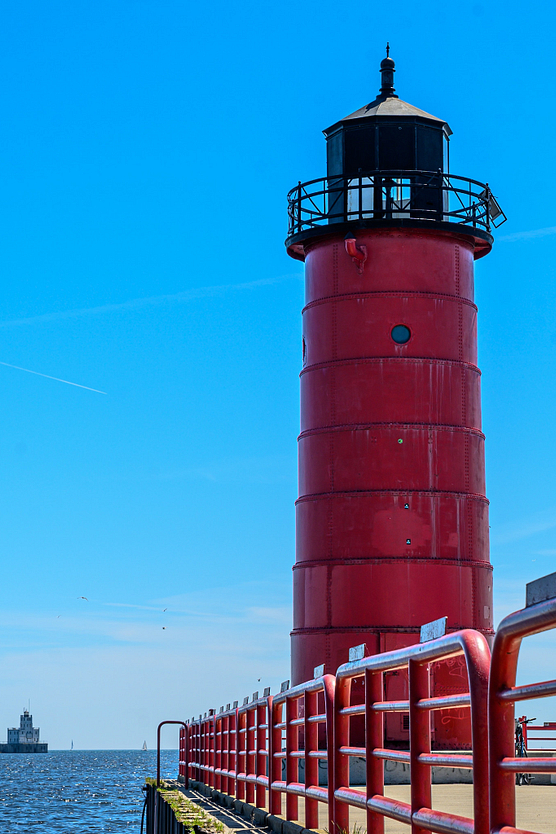 Red lighthouse against clear blue sky with sailboat and second lighthouse in Lake Michigan in the distance