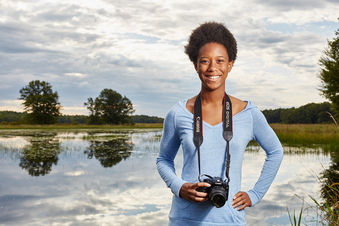 Photo of a smiling young woman with a camera around her neck, standing in the out-of-doors