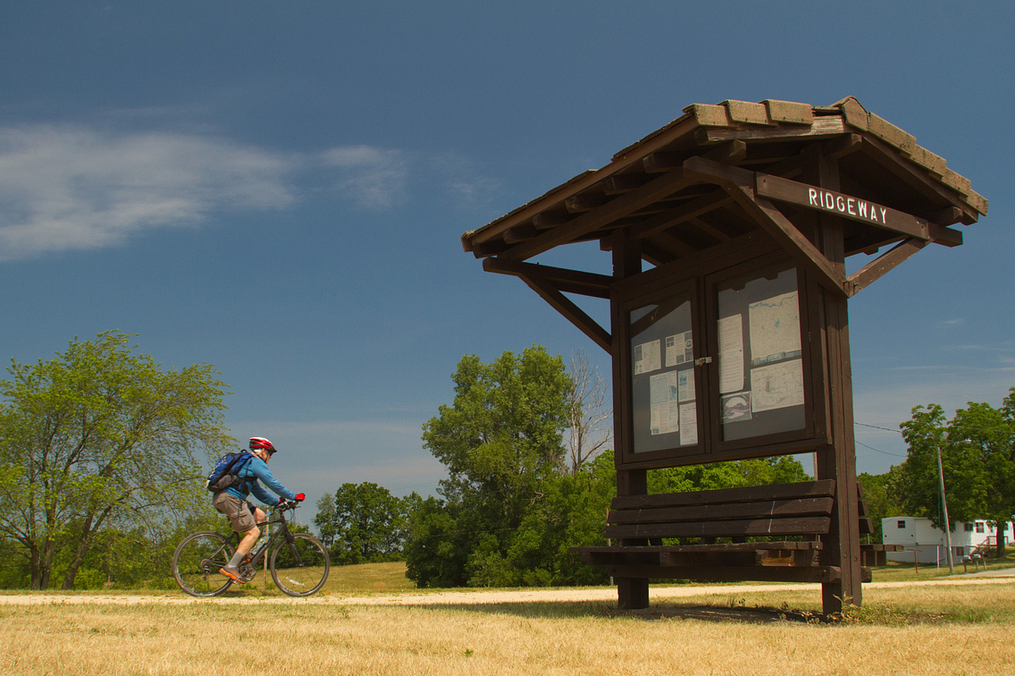 A bicyclist rides the Military Ridge State Trail near a kiosk that reads Ridgeway