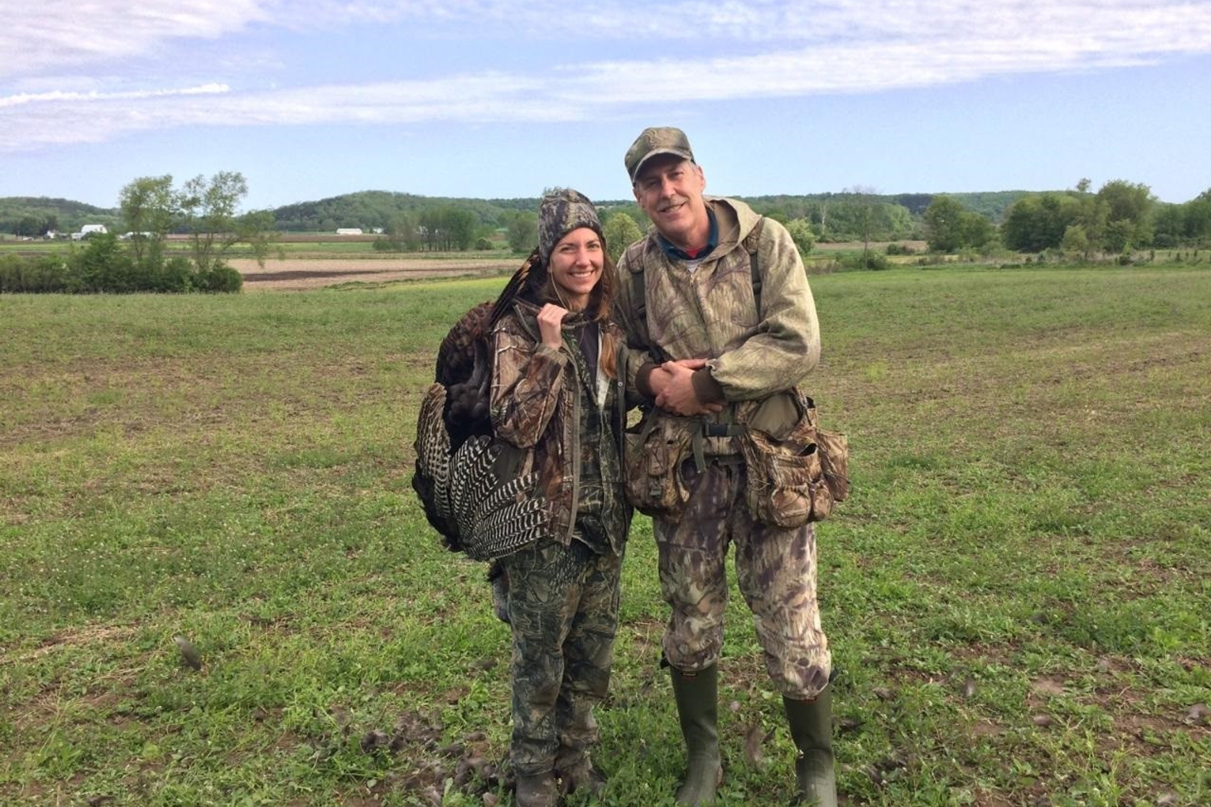 Two hunters in hunting clothing, standing in a field. Woman hunter on left carries a turkey she shot.